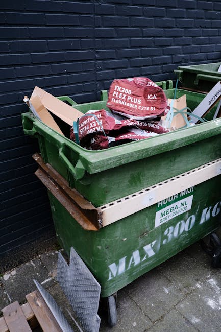 A green wheeled rubbish bin filled with multiple red and black bags of garden mulch or compost, placed on a pavement beside a dark blue brick wall. The bags have white and red text indicating they contain flexible garden products, positioned towards the top of the open bin. Beneath the bin, a white plastic container is partly visible, attached to the green bin. There is a wooden panel and some metal grating next to the wheel of the bin, and the ground consists of grey paving stones. The scene appears to be an outdoor residential or commercial area where waste or garden materials are temporarily stored, illustrating an example of private waste handling or alternative rubbish collection options. Rubbish Removal Docklands may be involved in the management or removal of such waste materials.