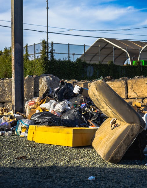 A narrow urban alleyway filled with accumulated waste and debris, featuring a large, grey, heavy-duty trash bag positioned in the foreground, with its textured fabric appearing taut and weathered. Behind it, a stack of flattened cardboard boxes, some with visible corrugation and torn edges, are piled haphazardly on a makeshift platform, indicating collection for disposal or recycling. To the left, a small black trash bin and a discarded white container lie on the uneven, slightly stained pavement, which shows signs of dirt, oil stains, and scattered litter. The alley is bordered by tall, weathered brick and metal-paneled walls, with graffiti visible on the right side, and a small set of windows and pipes attached to the buildings’ facades. Bare tree branches extend overhead against a pale, overcast sky, adding a stark, muted atmosphere typical of an environment awaiting rubbish collection. This scene exemplifies an informal rubbish accumulation area, representing a typical private waste handling point, which local services like Rubbish Removal Docklands might service as part of an alternative waste removal approach away from municipal collection.