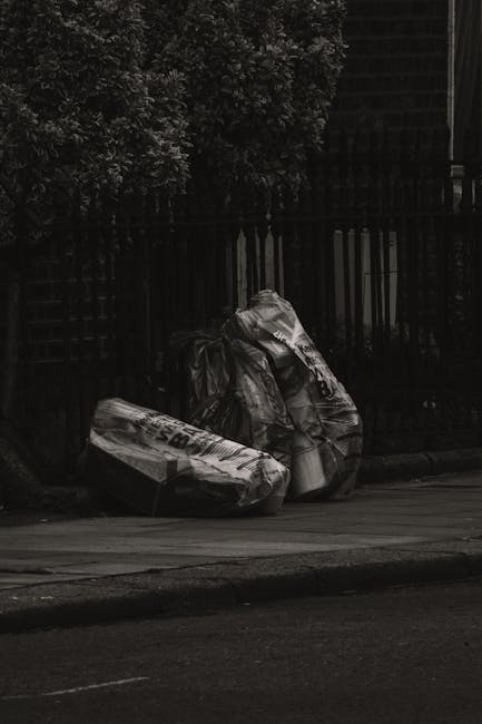 A pair of large black trash bags filled with waste materials are placed on the edge of a concrete pavement next to a dark metal fence with vertical bars. The bags appear to be made of thick, crinkled plastic and are slightly torn or stretched, indicating they are filled with bulky rubbish. The scene is outdoors, possibly on a street or alleyway, with a dense leafy tree visible behind the fence towards the top left corner, providing some natural backdrop. The overall lighting is subdued, suggesting overcast weather or low natural light, which emphasizes the dark tones and textures of the bags and the surrounding environment. This image, associated with rubbish removal services like those offered by Rubbish Removal Docklands, highlights typical informal waste disposal by residents or businesses in urban areas, contrasting with official waste collection methods and illustrating scenarios where private, independent rubbish handling might be necessary.