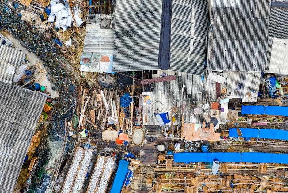 An aerial view of a densely packed outdoor area with various discarded materials and debris. The foreground features a mixture of scrap wood, pallets, and construction waste, some piled haphazardly and others arranged along wooden structures. Scattered across the scene are blue tarps covering sections of the debris, and the ground appears muddy and uneven. Slightly to the left, there are piles of plastic bottles and other mixed refuse, with some waste spilling onto adjacent pathways. In the background, several makeshift sheds or structures with corrugated metal roofs and walls are visible, with weathered and rusted finishes, indicating a site used for storage or informal waste management. The environment has a cluttered, utilitarian appearance typical of a private waste collection or scrap yard, reflecting an on-site removal or collection operation. This scene aligns with non-standard rubbish removal options, such as independent collection or private disposal, commonly undertaken outside local authority services. Rubbish Removal Docklands specializes in managing such diverse waste materials securely and professionally, supporting alternative waste handling methods.
