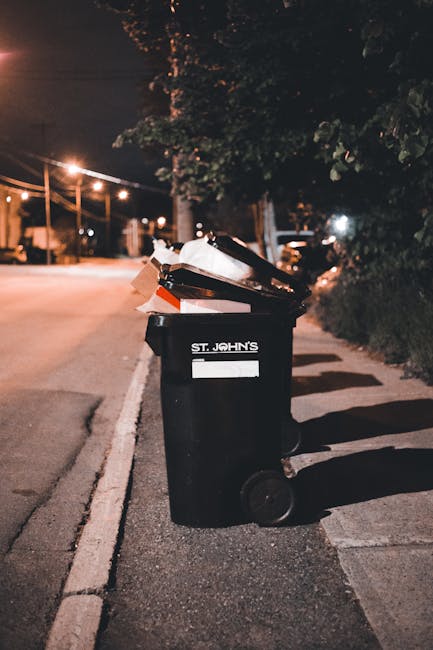 A black wheeled rubbish bin with a white label and the words 'ST. JOHN'S' printed on the front, placed on the pavement beside a curb. The bin is filled with various waste materials, including cardboard boxes, plastic packaging, and paper, some of which are overflowing and leaning out. It is positioned in an outdoor urban setting during nighttime, illuminated by nearby streetlights, casting shadows on the ground. In the background, there are trees, lamp posts, and parked cars visible along the quiet street. The scene suggests an instance of private waste disposal, where a bin like this might be used as an alternative to municipal rubbish collection, reflecting the type of disposal service that Rubbish Removal Docklands might support for local residents or businesses in the Docklands area.