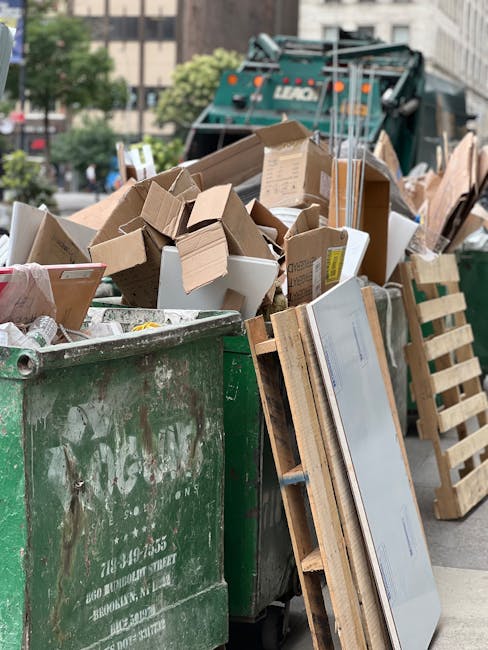 An aerial view of a densely packed outdoor area with various discarded materials and debris. The foreground features a mixture of scrap wood, pallets, and construction waste, some piled haphazardly and others arranged along wooden structures. Scattered across the scene are blue tarps covering sections of the debris, and the ground appears muddy and uneven. Slightly to the left, there are piles of plastic bottles and other mixed refuse, with some waste spilling onto adjacent pathways. In the background, several makeshift sheds or structures with corrugated metal roofs and walls are visible, with weathered and rusted finishes, indicating a site used for storage or informal waste management. The environment has a cluttered, utilitarian appearance typical of a private waste collection or scrap yard, reflecting an on-site removal or collection operation. This scene aligns with non-standard rubbish removal options, such as independent collection or private disposal, commonly undertaken outside local authority services. Rubbish Removal Docklands specializes in managing such diverse waste materials securely and professionally, supporting alternative waste handling methods.