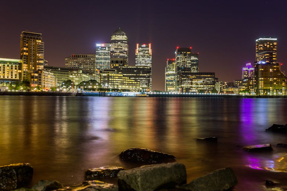 A nighttime view of a modern city skyline featuring illuminated high-rise buildings of various heights and architectural styles reflected in the calm water of a river or harbor in the foreground. The buildings display a mix of glass and concrete facades, with some structures highlighted by colorful lights in red, purple, and white, creating a vibrant urban atmosphere. The waterfront area includes some rocks and stones along the shoreline, partially submerged or resting on the edge of the water, providing a natural contrast to the illuminated cityscape. The scene captures the serene reflection of the city lights on the water, with gentle ripples slightly distorting the reflections. This urban waterfront setting, with its prominent skyline at night, aligns with themes of city-based waste management and the importance of effective rubbish removal services in densely populated areas, although no specific waste disposal elements are visible in the image.