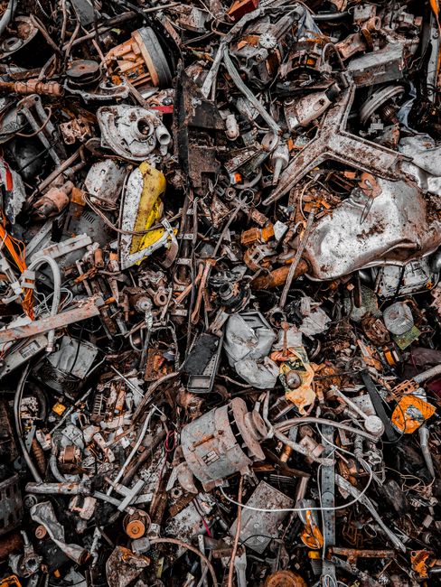A dense pile of discarded automotive parts and machinery components, including rusted metal engine blocks, gears, panels, and various mechanical parts, all piled together in a scrapyard setting. The metal surfaces exhibit corrosion, discoloration, and worn textures in shades of brown, black, and silver, with some parts covered in dirt and grease. The scene shows an assortment of broken or decommissioned vehicle elements, with some wires and cables intertwined amongst the debris. The background reveals additional scrap metal and tools, suggesting an environment of industrial dismantling or waste disposal. This image illustrates the type of heavy steel and mechanical waste materials that can be managed through independent rubbish removal services like those offered by Rubbish Removal Docklands, which handle such bulky and complex waste items as part of their on-site clearance solutions.