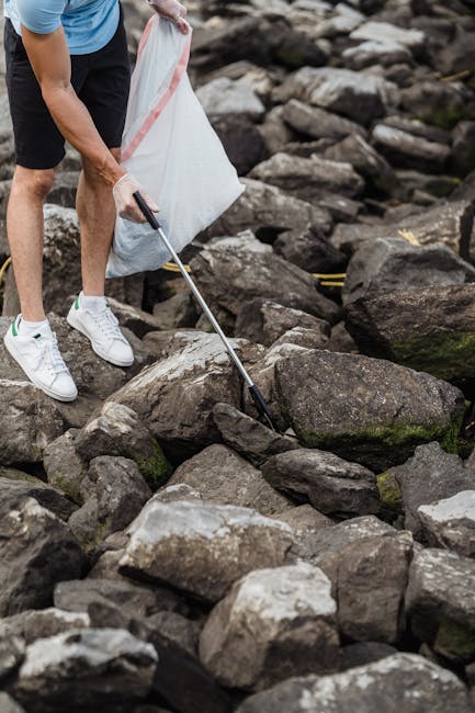 A person wearing a light blue t-shirt, dark shorts, and white sneakers is using a litter picker to collect small pieces of rubbish placed among large, irregularly shaped rocks on a rocky outdoor surface. The rocks vary in shades of dark gray, brown, and green, with some moss or algae growth visible on their surfaces. The individual is holding a white plastic bag with a red stripe in one hand, positioned close to the rocks, indicating an activity of cleaning or waste collection. This scene suggests a focus on environmental maintenance or outdoor waste removal, paralleling the services provided by Rubbish Removal Docklands that specialize in efficient, private waste collection and on-site clearance of debris, including rubbish accumulated in natural or urban environments. The natural setting, combined with the use of tools for litter picking, highlights a responsible approach to waste management outside traditional waste disposal sites, supporting eco-friendly practices and alternative rubbish removal methods.